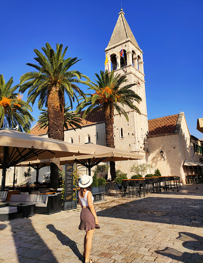 Qué ver en Trogir: Yo frente a la Iglesia y Monasterio de Santo Domingo con unas palmeras delante y una terraza con toldos