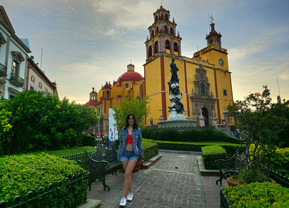 En la Basílica de Nuestra Señora de Guanajuato con sus colores amarillo y rojo al fondo de la imagen, frente al jardín que se encuentra en la plaza con una banca de cada lado - Qué ver en Guanajuato