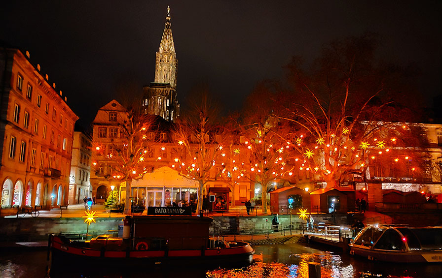Árboles con estrellas y luces y la catedral de fondo. Y dos barquitos en el río - Qué ver en Estrasburgo