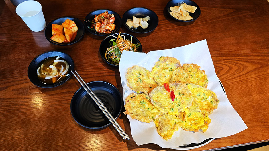 Plato de Pajeon acompañado de banchan en boles negros y unos palillos a la izquierda - Qué comer en Corea del Sur