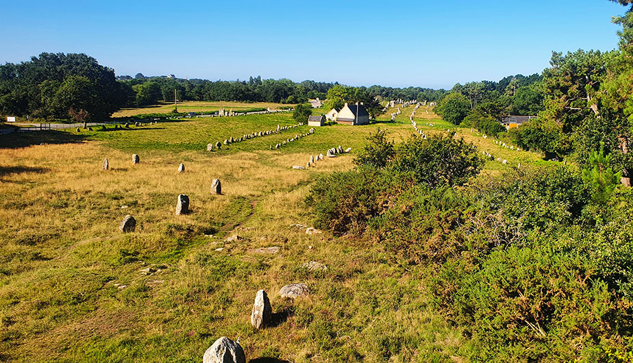 Línea de piedras en un enormes prado con unas pequeñas casas al fondo - Ruta por la Bretaña Francesa