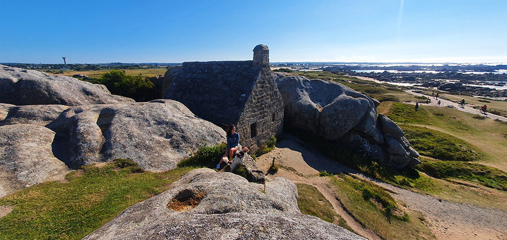 Sentada en una roca con una ermita de piedra detrás y más rocas enormes y el mar al fondo - Ruta por la Bretaña Francesa