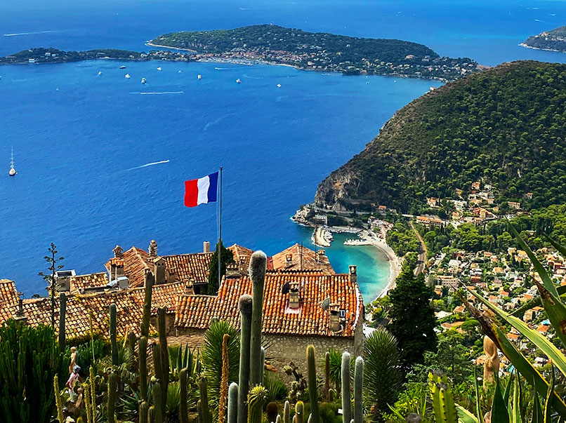 Vistas de la costa, con algunas suculentas de frente, con algunas casas debajo y la bandera de Francia - Qué ver en Èze