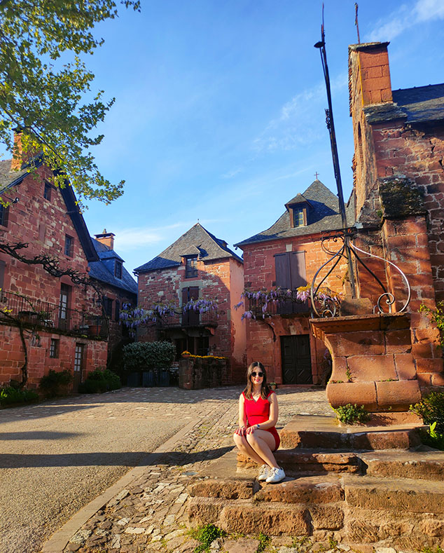 Sentada sobre unas escaleras con una cruz detrás y casas rojas con flores moradas al fondo en Collonges-la-Rouge