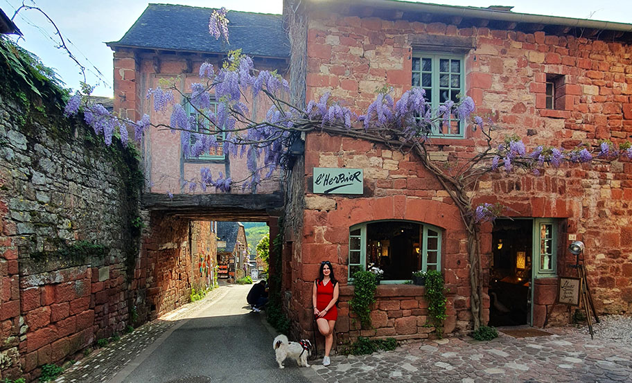 Apoyada junto con mi perro en una pared rojiza con ventana en verde y un pasadizo a la izquierda con flores moradas en Collonges-la-Rouge