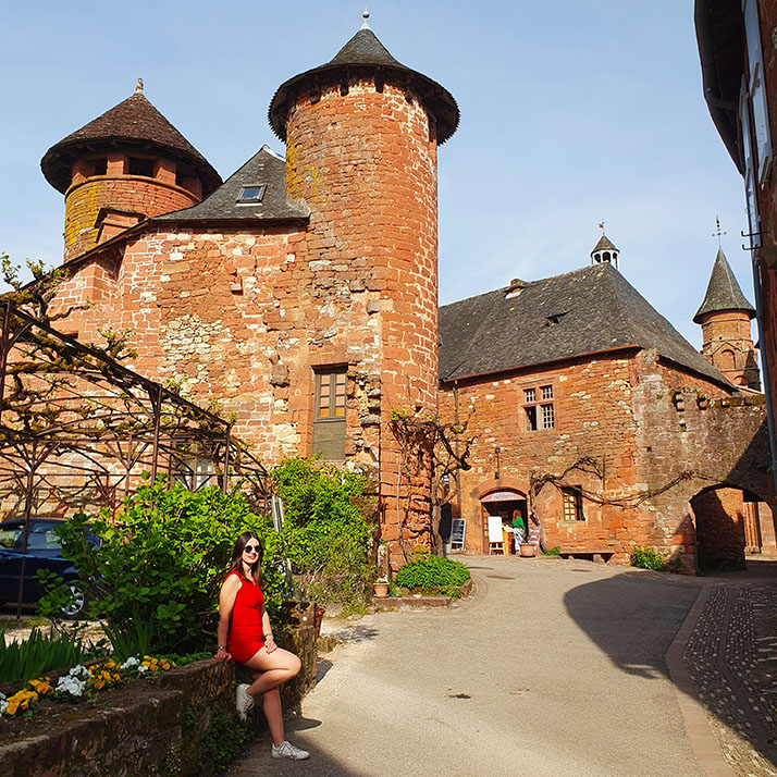 Apoyada en una zona con flores y pequeños arbustos y con edificios rojos al fondo con torres en Collonges-la-Rouge