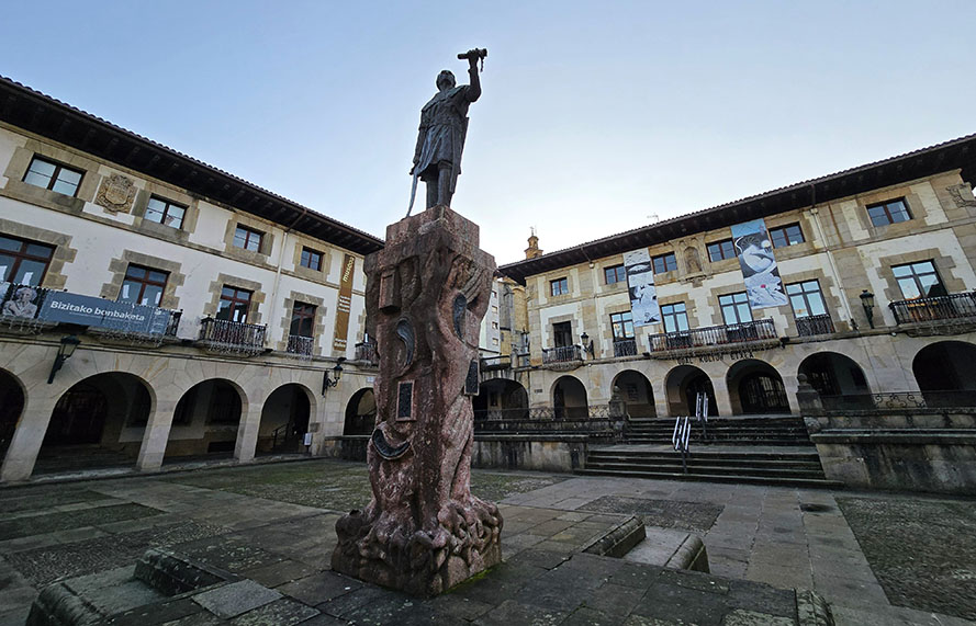 Escultura en medio de una palza con edificios atrás y pórticos - Qué ver en Gernika
