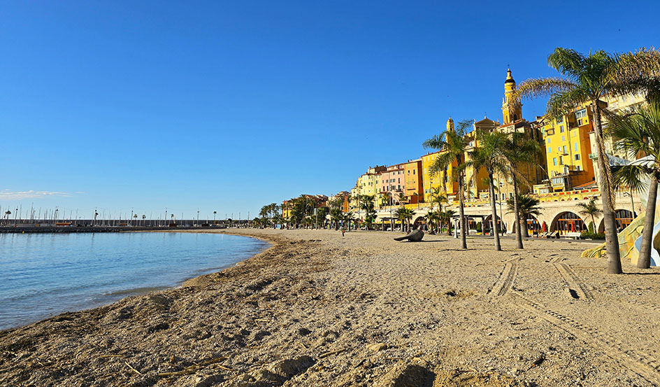Playa con palmeras y casas de colores amarillos, naranjas y rosas en un costado - Qué ver en Menton