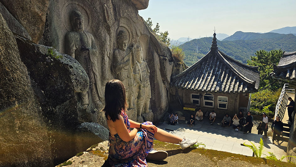 Sentada observando un pequeño templo, con budas tallados en la pared de roca en el lado izquierdo y mucha vegetación al fondo - Ruta por Corea del Sur