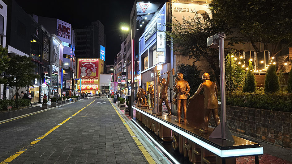 Calle de noche con figuras de oro a la derecha y edificios con neones al fondo - Dónde alojarse en Busan
