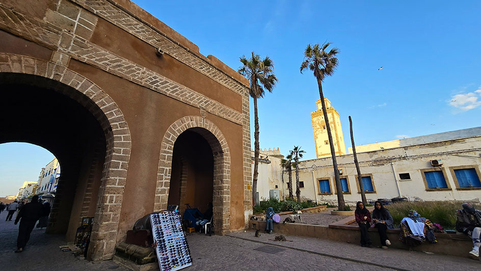 Puerta con arcos, con un puesto de gafas apoyado, chicas sentadas a la derecha y el minarete de la mezquita al fondo - Qué ver en Essaouira