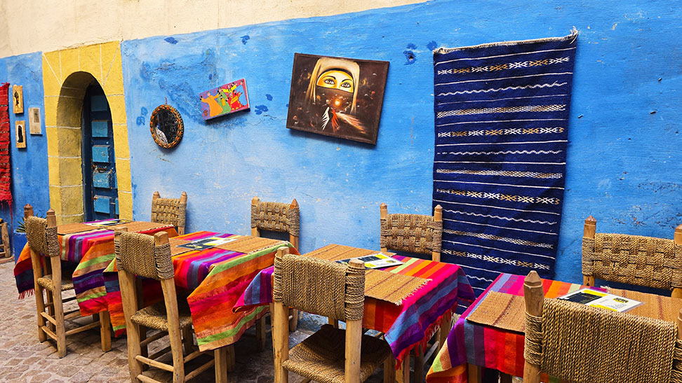 Pared azul con varias mesas y sillas con mantel lle o de colores - Preparativos para viajar a Marruecos por libre
