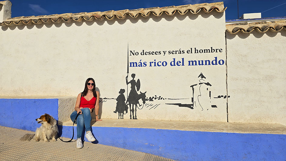 Sentada con mi perro juno a un mural de Don Quijote y Sancho Panza con un molino y una frase "No desees y serás el hombre más rico del mundo" - Qué ver en Campo de Criptana