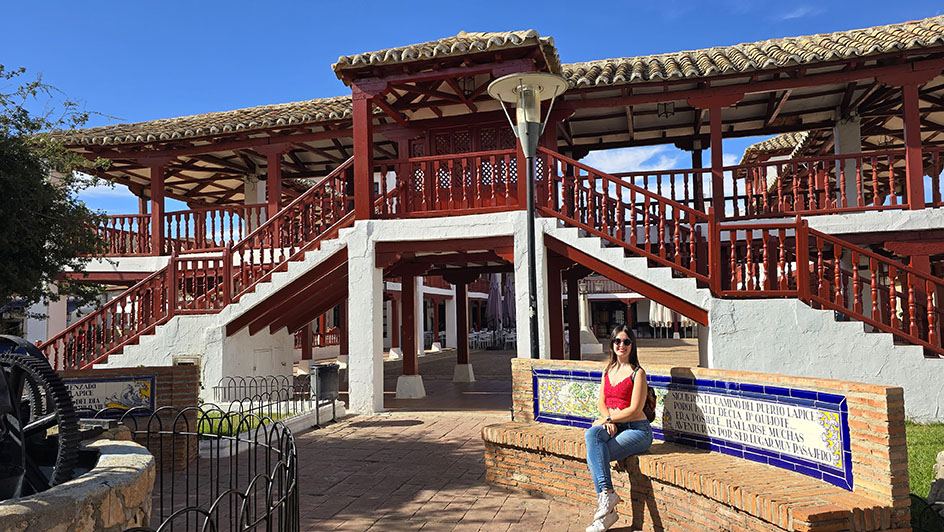 Sentada en un banco de ladrillo con azulejos y detrás una plaza de dos pisos con balcones de madera rojos - Qué ver en Puerto Lápice
