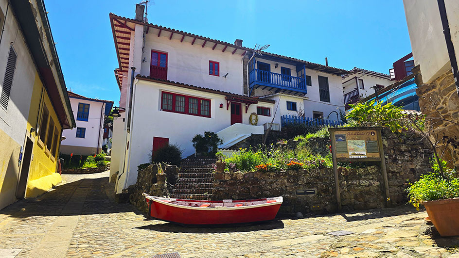 Calles empedradas, una barca roja frente a unas casas blancas escalonadas con detalles de madera pintadas en rojo y azul. - Qué ver en Tazones