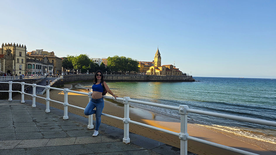 Apoyada sobre las barandillas blancas frente a una playa en el paseo marítimo y una iglesia al fondo - Qué ver en Gijón