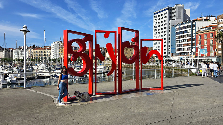 Posando junto a mi perro con las letras de Gijón de color rojo frente al puerto depostivo lleno de barcos - Qué ver en Gijón