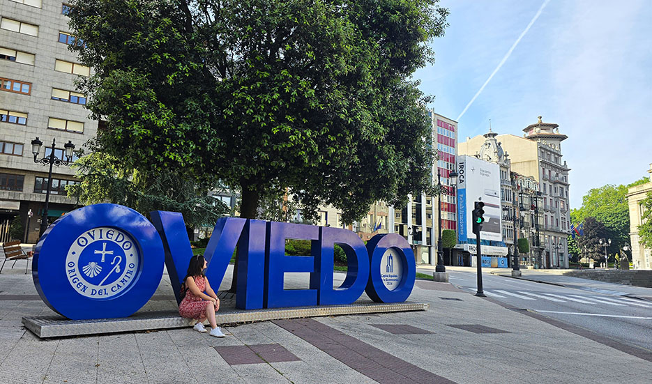 Sentada junto a las Letras de Oviedo de color azul con un árbol al fondo - Qué ver en Oviedo