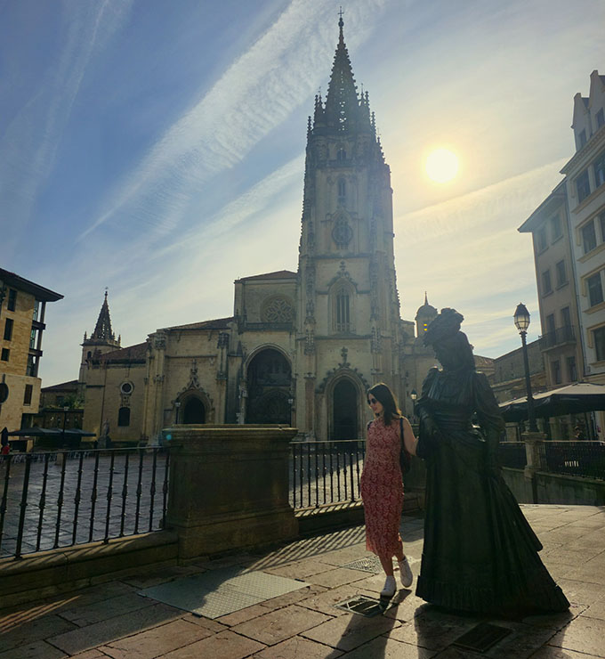 Apoyada mirabdo al suelo junto a la escultura de una mujer en una plaza con una catedral al fondo - Qué ver en Oviedo